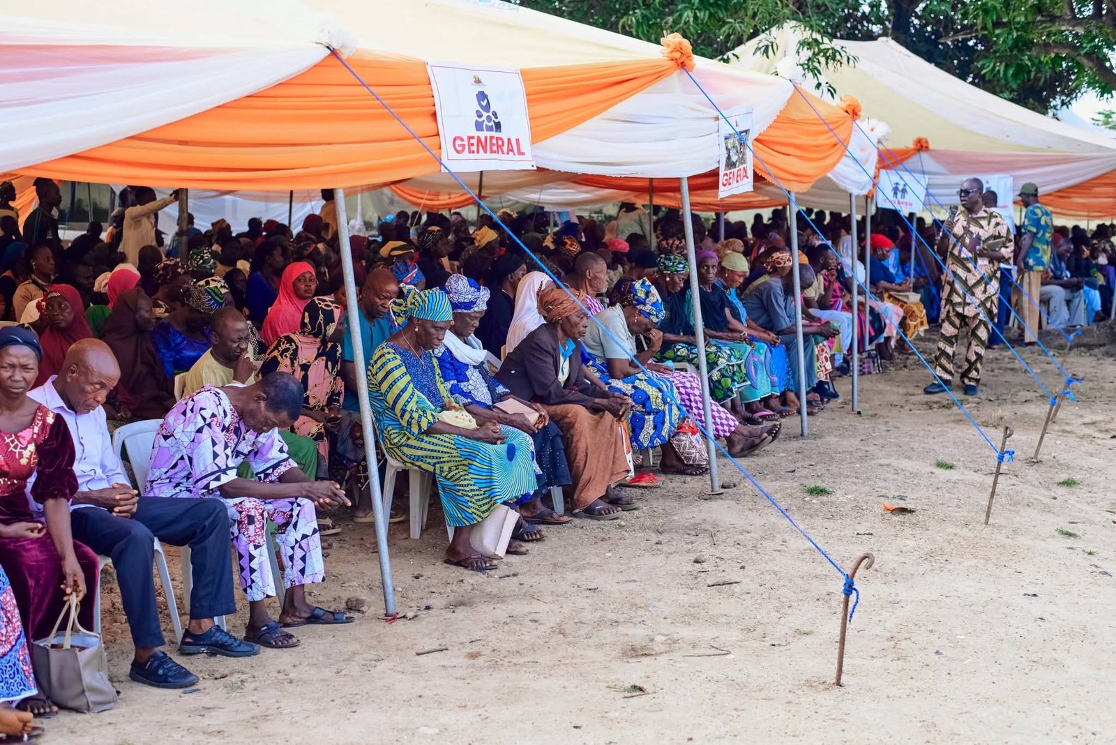 Community members seated beneath a large outreach canopy.
